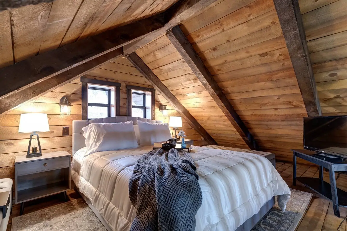 Cozy attic bedroom with wood-paneled walls and ceiling. Features a large bed with white bedding, two bedside tables with lamps, and a small TV on a stand.