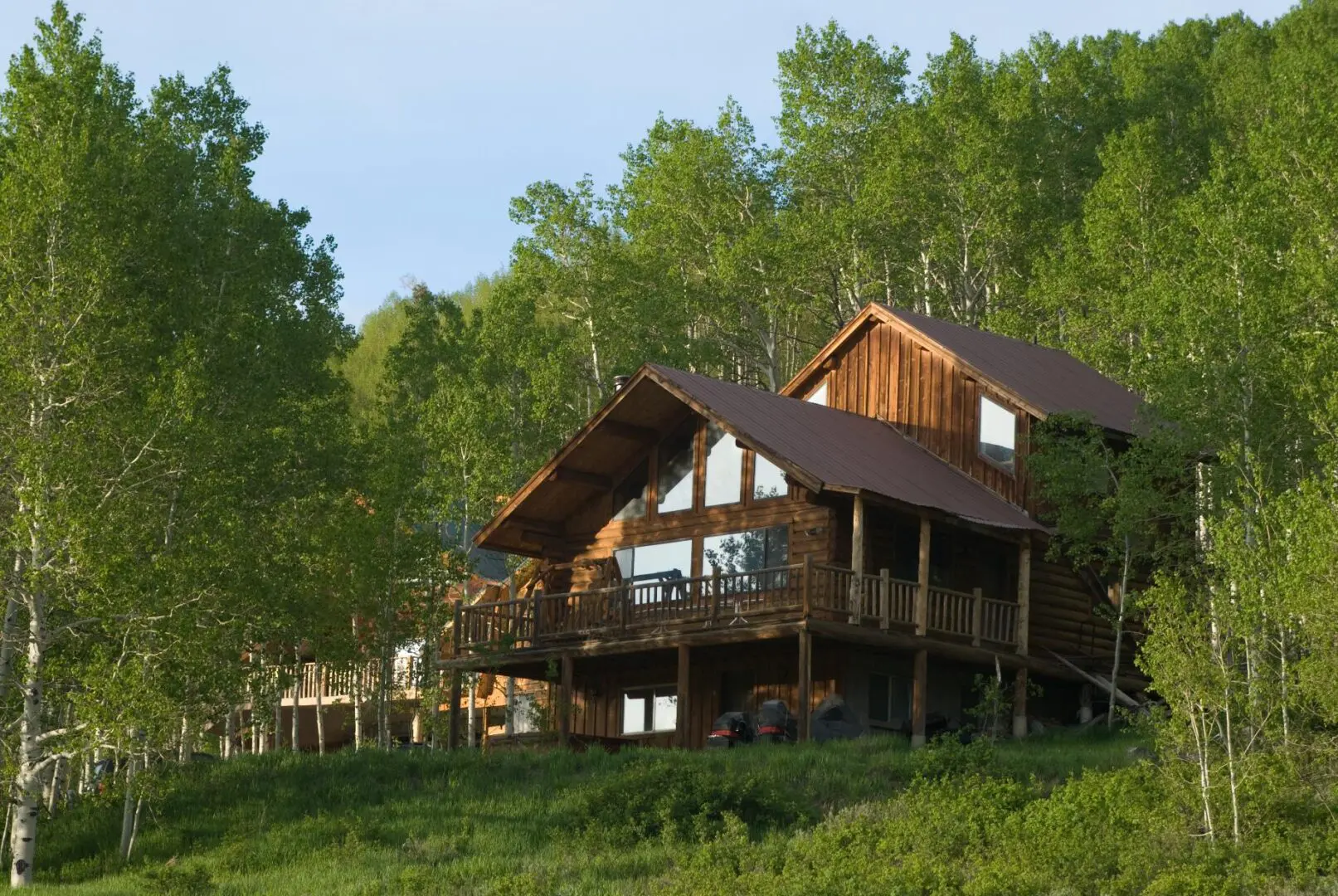 Wooden cabin with a sloped metal roof surrounded by lush green trees on a sunny day. The cabin has large windows and a wraparound porch.