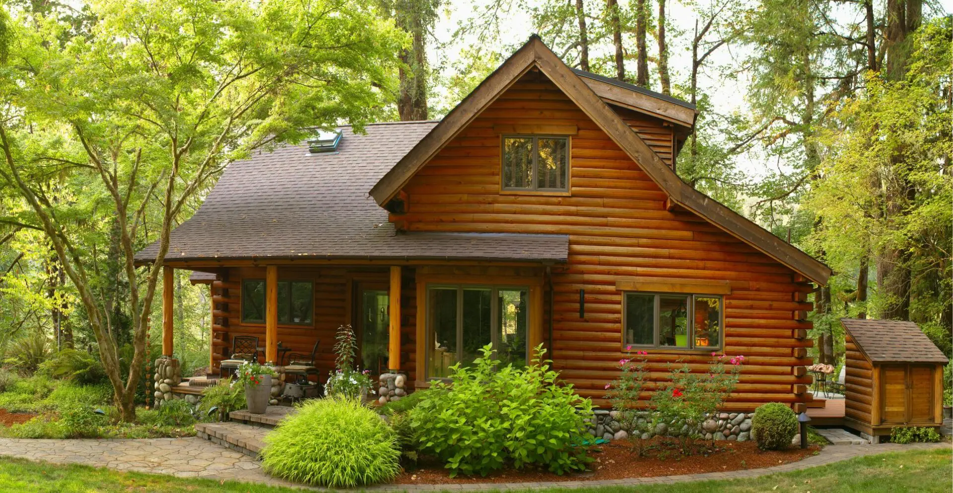 A wooden cabin surrounded by trees and greenery, with a small front porch and shrubs.