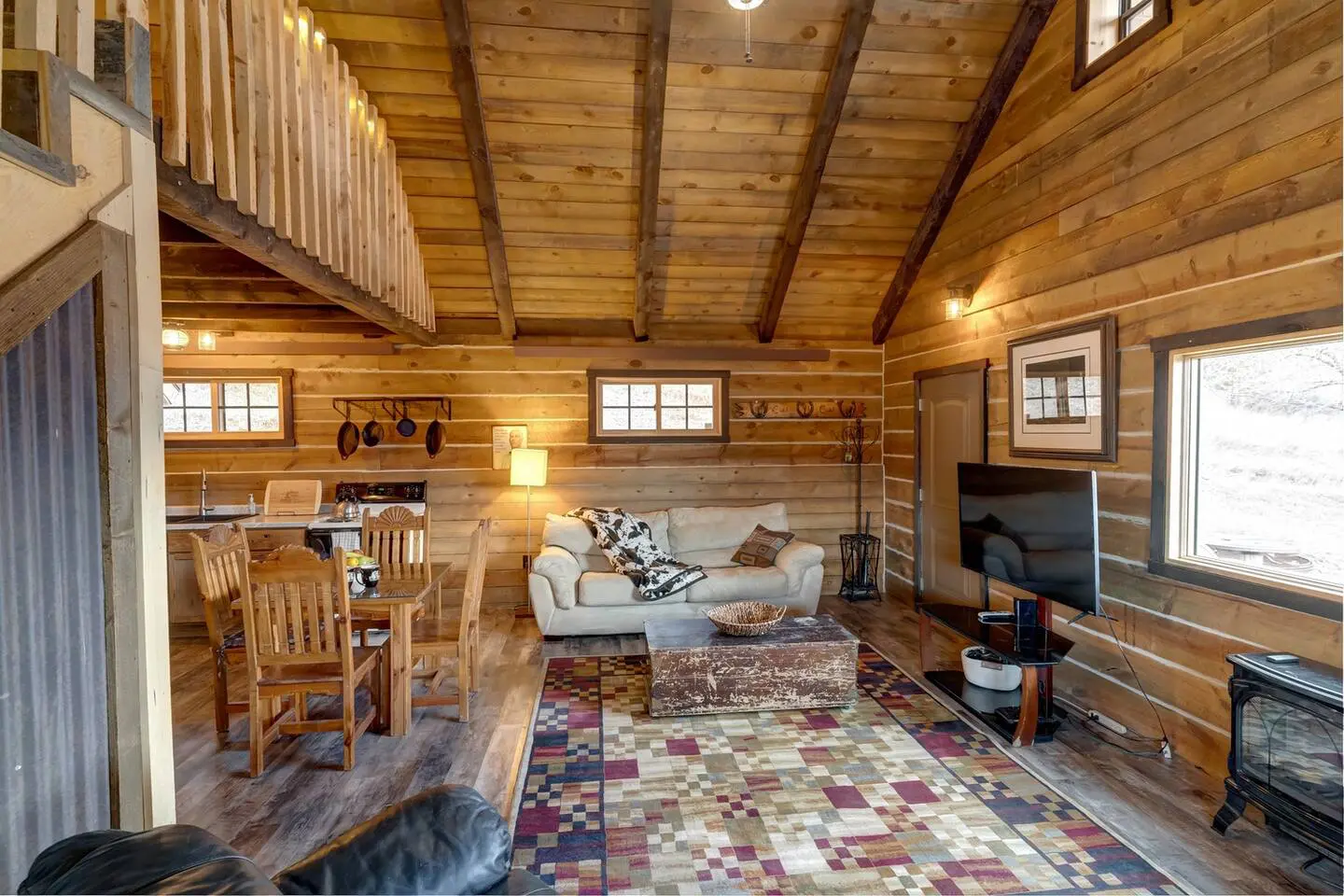 Cozy cabin interior with wooden walls, a sofa, dining table, TV, and colorful rug. Loft area above dining space. Warm lighting and decor create a rustic atmosphere.