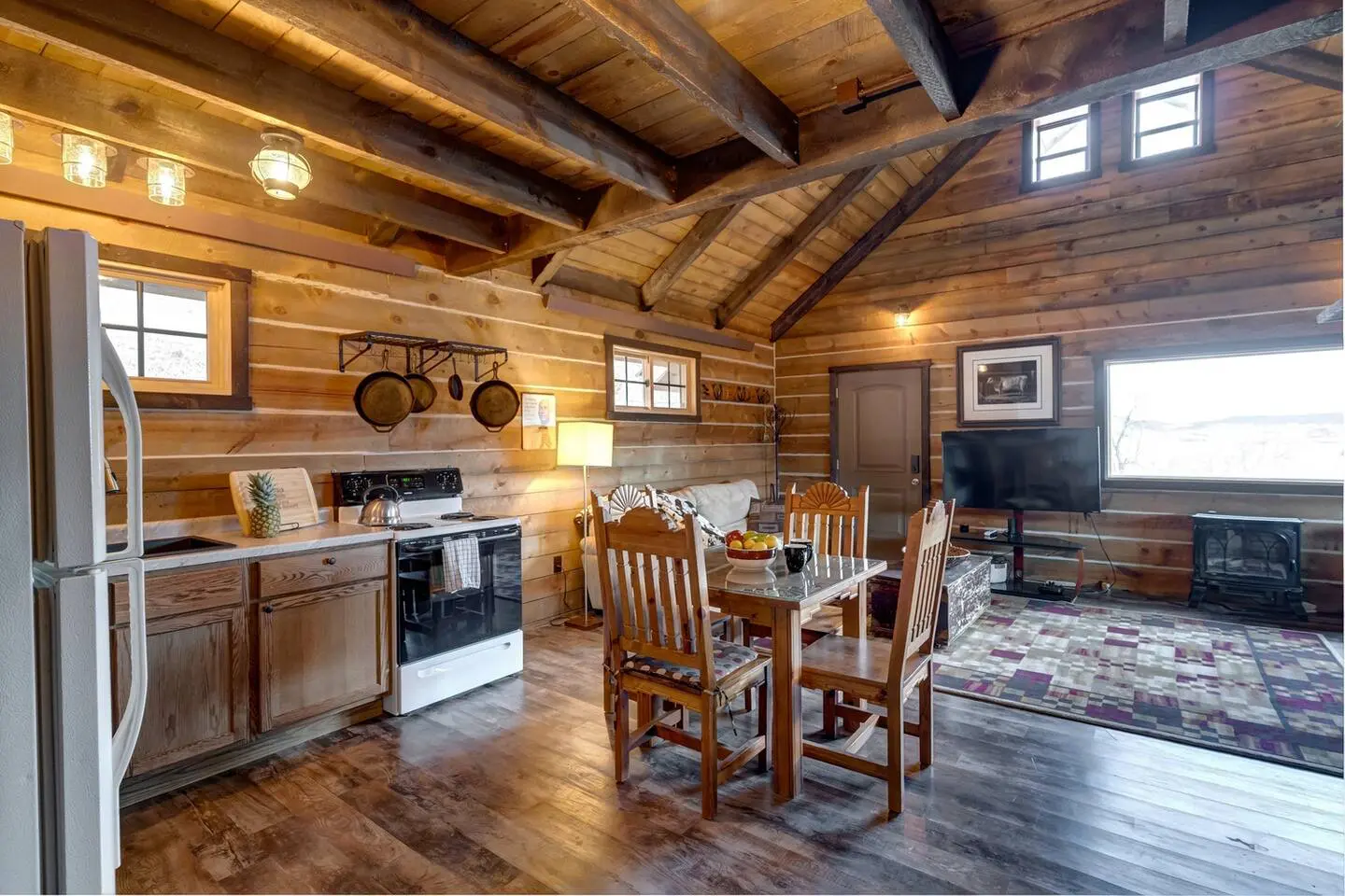 A rustic kitchen with wooden walls and beams features a dining table with chairs, a stove, and a refrigerator. A small living area with a TV is visible in the background.