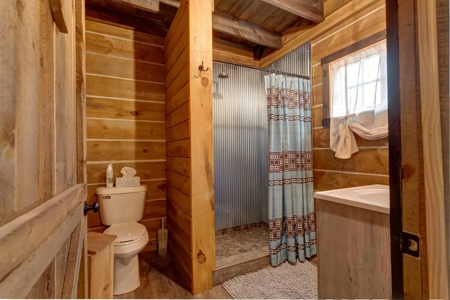 Rustic bathroom with wooden walls, a shower stall with a patterned curtain, a toilet, and a sink with a window above it. Natural light filters in through the window.