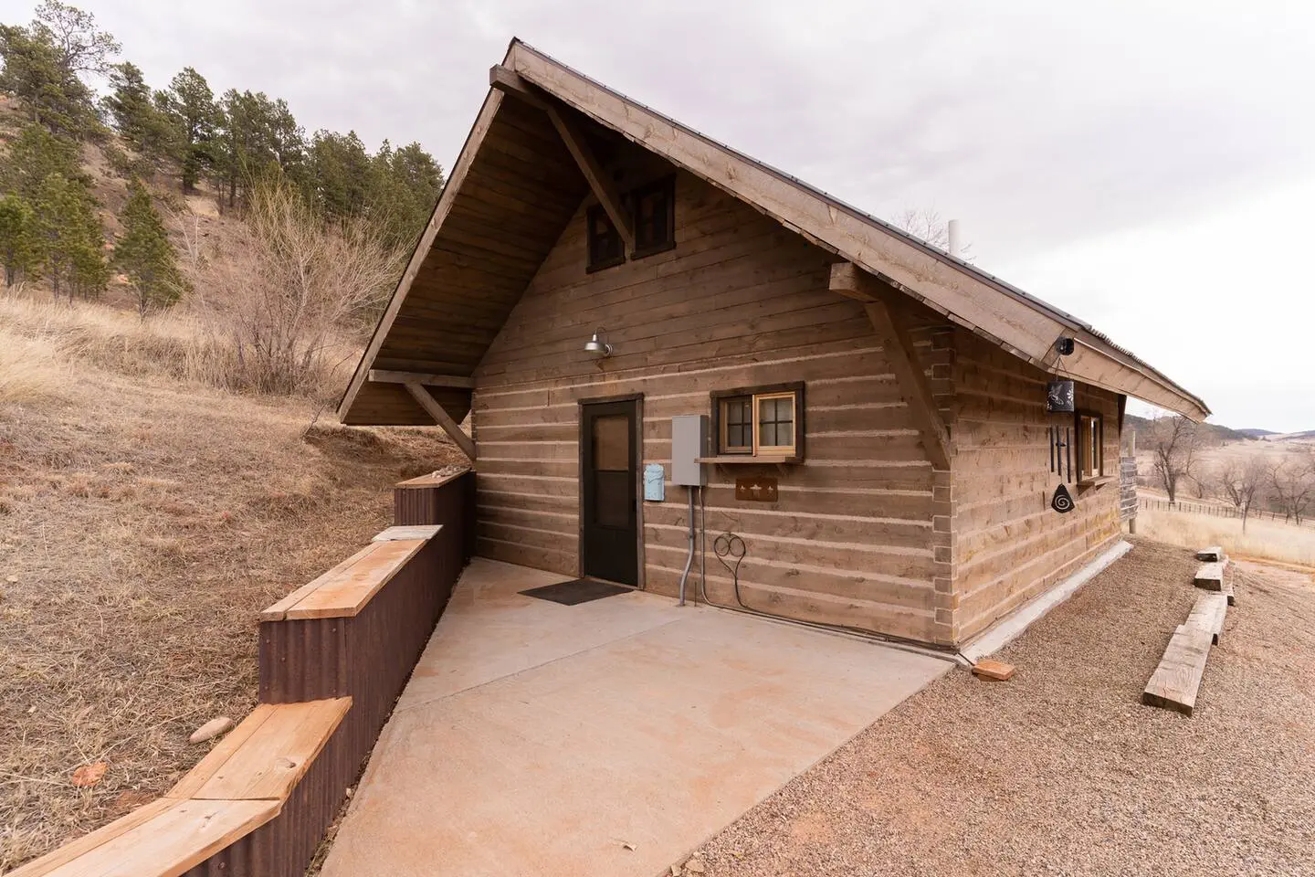 Wooden cabin with a sloped roof, set on a hillside with a concrete path leading to a door. Surrounded by dry grass and trees under a cloudy sky.
